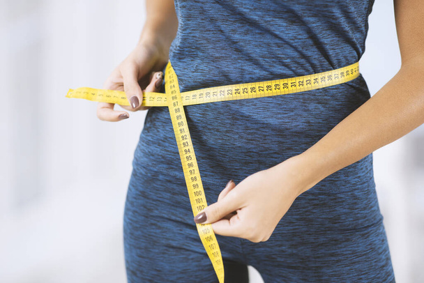 Conceito de perda de peso. Mulher magro em sportswear medindo sua cintura com fita métrica dentro de casa, vista close-up
 - Foto, Imagem