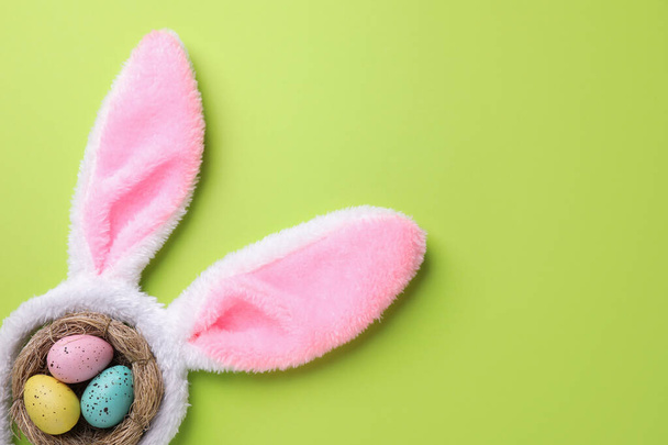 Headband with Easter bunny ears and dyed eggs in nest on green background, flat lay. Space for text - Foto, afbeelding