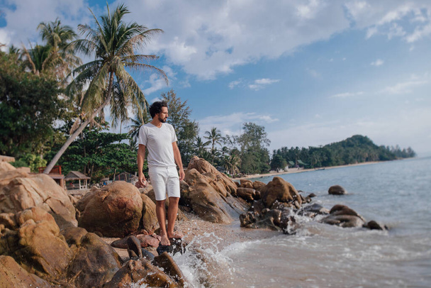 ein Mann an der tropischen Küste in Steinen am Meer. Ein junger gutaussehender Mann in weißer Kleidung steht an einem felsigen Strand vor dem Hintergrund von Palmen und dem Meer. Gesamtplan. - Foto, Bild