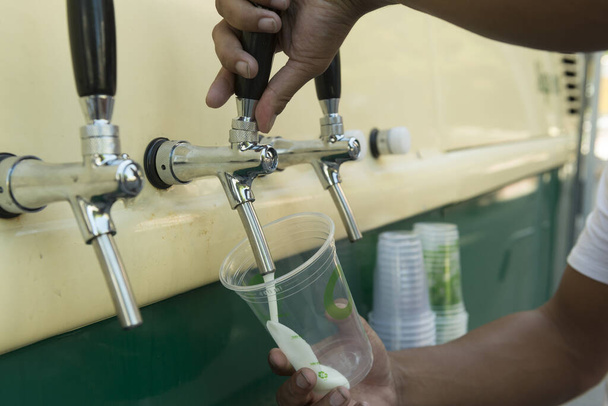 Μπάρμαν ρίχνει από τη βρύση φρέσκια μπύρα στο πλαστικό κύπελλο. Χέρια μπάρμαν γεμίζουν ποτήρια μπύρας από μια βρύση μπαρ. Χέρι μπάρμαν ρίχνει μια μπύρα Πίλσεν στη βρύση. Εμπόριο οδών και τοπικός πολιτισμός. - Φωτογραφία, εικόνα