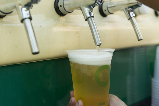 Μπάρμαν ρίχνει από τη βρύση φρέσκια μπύρα στο πλαστικό κύπελλο. Χέρια μπάρμαν γεμίζουν ποτήρια μπύρας από μια βρύση μπαρ. Χέρι μπάρμαν ρίχνει μια μπύρα Πίλσεν στη βρύση. Εμπόριο οδών και τοπικός πολιτισμός. - Φωτογραφία, εικόνα
