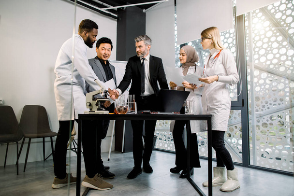 Interracial group of business men and doctors scientists team meeting in modern lab or office, standing at the table with laptop, microscope and glasswear. Handshaking of Caucasian and African men - Photo, image