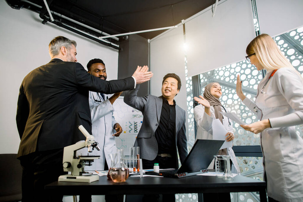 Happy multiethnical business scientists doctors team making high five each other while standing at the table with lab equipment. Scientific success, new research, achievement, team work concept - Photo, image