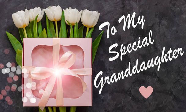 Στο ειδικό μήνυμα για την εγγονή μου. Λευκά λουλούδια τουλίπας και ροζ κουτί δώρου με κορδέλα φιόγκο σε σκούρο φόντο επίπεδη θέσει. Χρόνια πολλά λουλούδι μπουκέτο ευχετήρια κάρτα. Γυναικείο blog banner top view. - Φωτογραφία, εικόνα