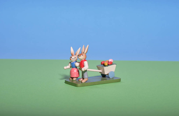 En un prado verde bajo un cielo azul, dos conejos de Pascua tiran de un vagón con huevos coloreados
. - Foto, imagen
