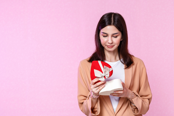 a young girl in a beige jacket with a gift box in her hands. surprised and happy with the gift. on a pink isolated background. - Foto, afbeelding