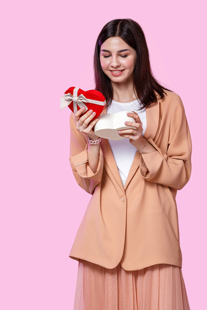 молода дівчина в бежевій куртці з подарунковою коробкою в руках. здивований і щасливий подарунок. на рожевому ізольованому фоні
. - Фото, зображення