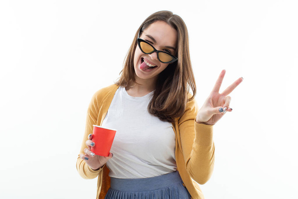 Brunette in a yellow sweater drinks coffee from a red cup and shows tongue - Photo, Image