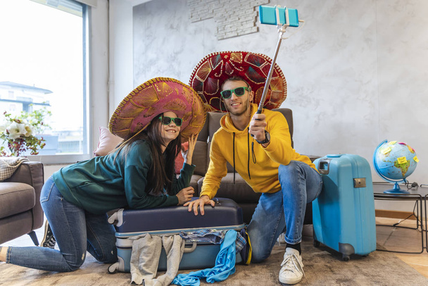 young couple sitting on the floor at home takes a selfie with smartphone wearing colorful sombrero before leaving for the vacations - Foto, Imagem