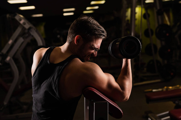 A handsome man with glasses doing an exercise for biceps with a barbell. The guy is engaged in bodybuilding. Trainer in the gym with muscular arms. - Foto, afbeelding