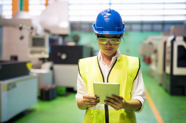 Tailliertes Porträt einer Arbeiterin mit Hut und Schutzbrille, die mit einem Tablet die Applikation von Unternehmen überprüft, um automatisierte Robotermaschinen in der Fabrik zu überprüfen. Fertigungsindustrie mit Technologie. - Foto, Bild