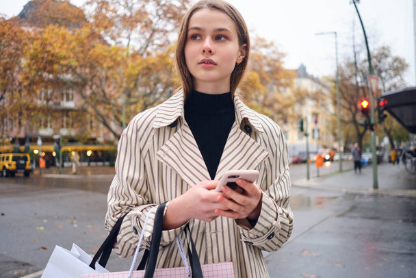 Довольно случайная девушка задумчиво ходить по уютной улице города с сумками и смартфоном
 - Фото, изображение