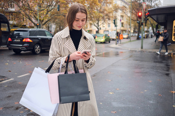 Atraktivní stylová dívka s nákupními taškami šťastně pomocí smartphone stojící na útulné městské ulici - Fotografie, Obrázek