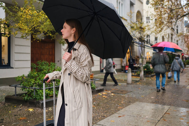 Mooi stijlvol meisje wandelen rond regenachtige stad straat met paraplu en koffer - Foto, afbeelding