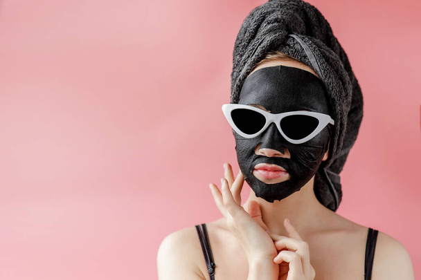Young woman in glasses appling black cosmetic fabric facial mask on pink background. Face peeling mask with charcoal, spa beauty treatment, skincare, cosmetology. Close up. - 写真・画像