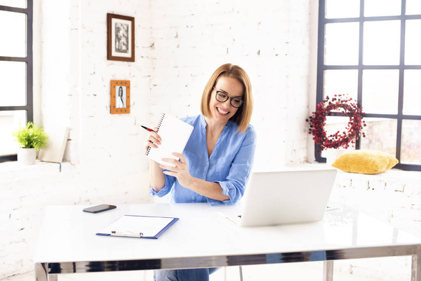 Zdjęcie atrakcyjnej dojrzałej bizneswoman siedzącej za biurkiem za notatnikiem i wykonującej papierkową robotę podczas pracy w biurze.  - Zdjęcie, obraz