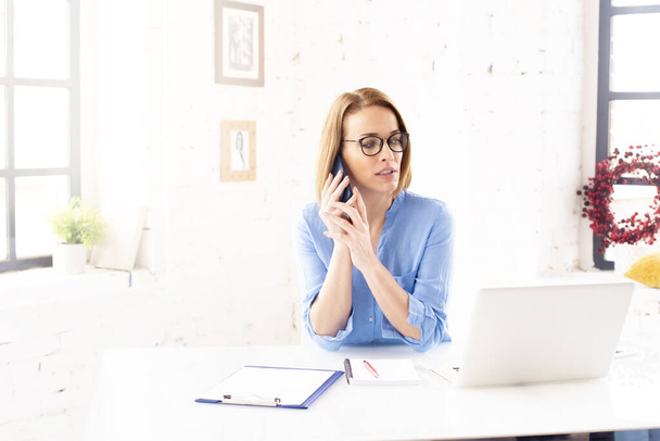 Знімок привабливої зрілої бізнес-леді, що сидить за ноутбуком і робить бізнес дзвінок під час роботи в Інтернеті
.  - Фото, зображення