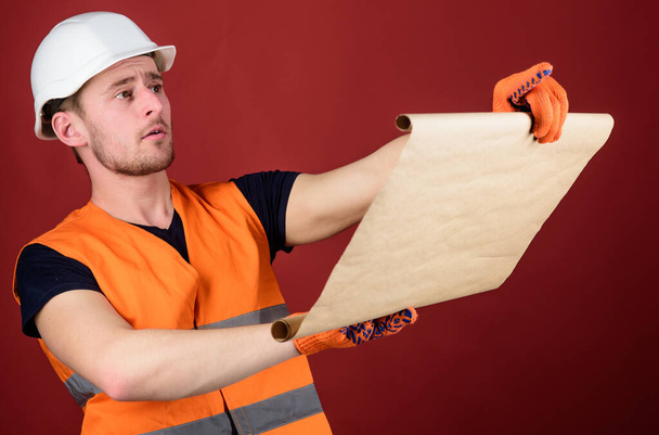 Engineer, architect, builder on strict face holds old blueprint in hands, supervises construction site. Man in helmet, hard hat holds building plan, controls works, red background. Foreman concept. - Photo, image