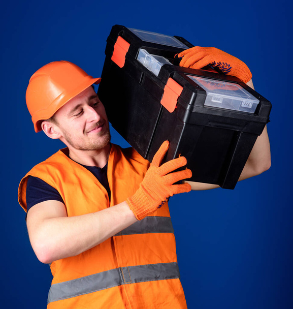 Worker, repairer, repairman, strong builder on cheerful smiling face carries toolbox on shoulder, ready to work. Repair service concept. Man in helmet, hard hat holds toolbox, blue background. - Valokuva, kuva