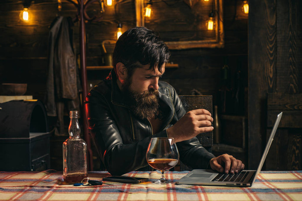Stylish handsome bearded man enjoying whiskey and doing his business at laptop. Elegant man in leather jacket drinking a whiskey in bar. Brutal hipster man drinking alcohol. - Zdjęcie, obraz
