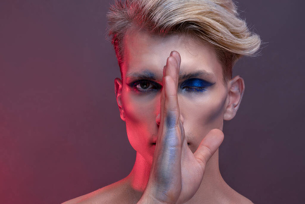 Portrait of a young man with professional beauty makeup and blond flying hair on a red background dividing face by hand in two halves, beautiful eyes, natural clean skin, hair care concept - 写真・画像