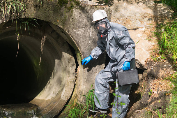 an environmental laboratory specialist in a protective suit and mask holds a case with a mobile laboratory in his hands and goes to the epicenter of the flow of chemicals - Foto, Imagem