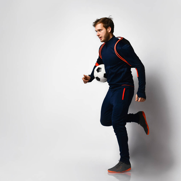 Giocatore di calcio in tuta blu, scarpe da ginnastica e zaino. Saltando su, sorridendo, tenendo la palla, posando isolato sul bianco. A tutta lunghezza
 - Foto, immagini