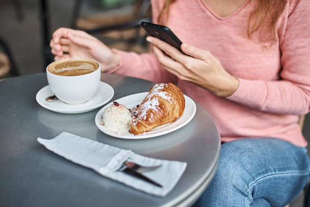 Woman have breakfast in caffe and using smartphone. Girl chatting and using internet with phone during coffee break with croissant - Valokuva, kuva