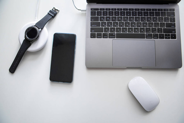 白いテーブルの上のワイヤレス充電器で携帯電話のコンピュータのマウスとスマートウォッチ充電とノートパソコン - 写真・画像