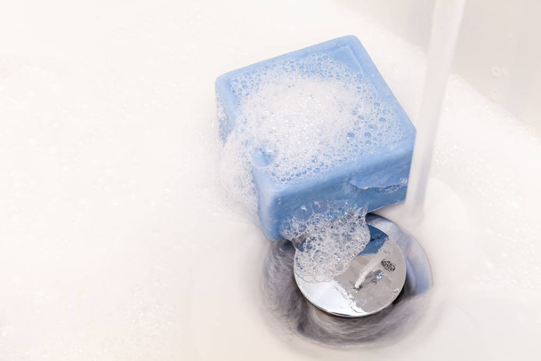 Bar of square blue soap and suds in a white sink with chrome plug and runnning water tap. Copy space - Fotografie, Obrázek