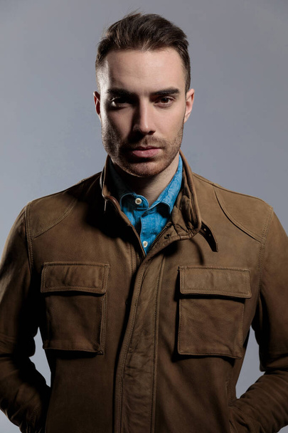 Modelo de moda guapo mirando hacia adelante con ambas manos en los bolsillos mientras usa chaqueta de cuero, de pie sobre fondo de pantalla gris
 - Foto, imagen