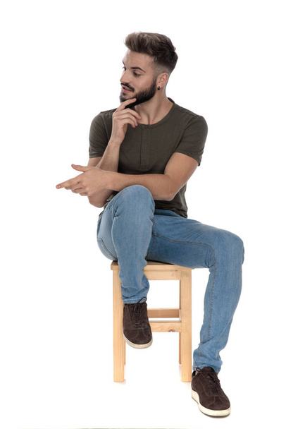 Jovem homem casual sorrindo e alcançando um aperto de mão enquanto sentado em uma cadeira no fundo do estúdio branco
 - Foto, Imagem