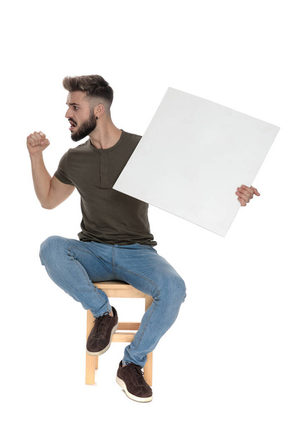 Angry casual man shouting and gesturing while holding a blank billboard, sitting on a chair on white studio background - Фото, изображение