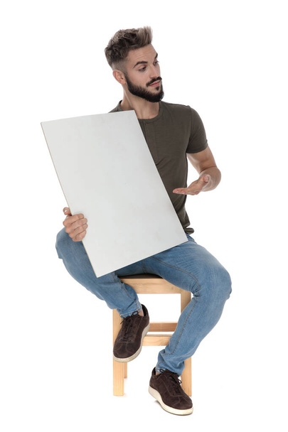 Friendly casual man presenting a blank billboard, sitting on a chair on white studio background - Photo, image
