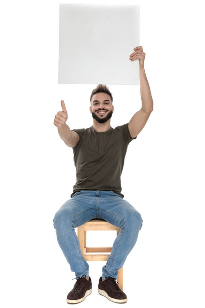 Positive casual man holding a blank billboard and gesturing ok, sitting on a chair on white studio background - Foto, Imagen