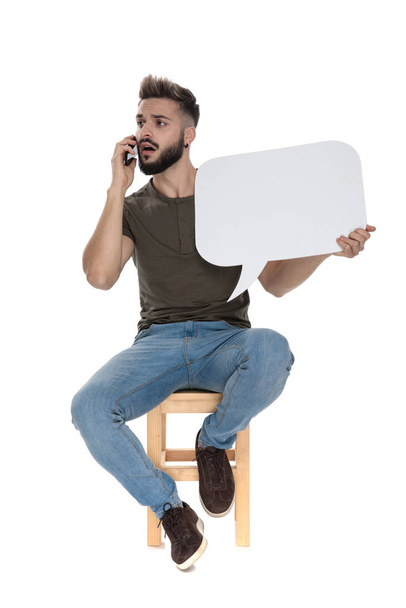 Worried casual man talking on phone and holding a blank speech bubble, sitting on a chair on white studio background - Photo, Image