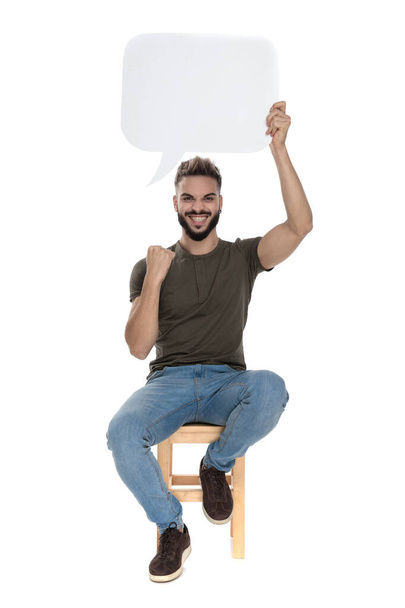 Happy casual man celebrating and holding blank speech bubble, sitting on a chair on white studio background - Φωτογραφία, εικόνα