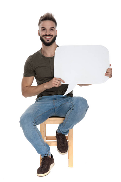 Homme occasionnel joyeux tenant une bulle de discours vierge et souriant, assis sur une chaise sur fond de studio blanc
 - Photo, image