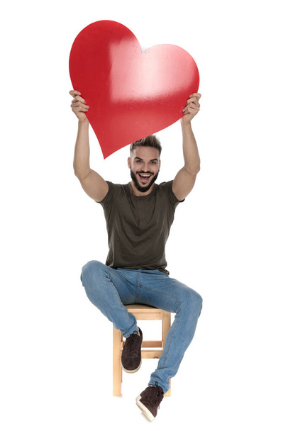 Cheerful casual man celebrating and holding a heart while sitting on a chair on white studio background - Фото, изображение