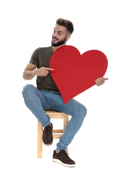 Hombre casual positivo apuntando y sosteniendo un corazón mientras está sentado en una silla sobre fondo blanco del estudio
 - Foto, Imagen
