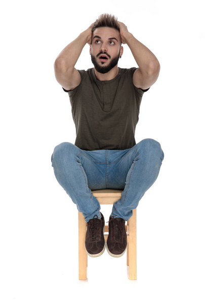 Amazed casual man holding his hands on his head and gasping while looking up and sitting on a chair on white studio background - Photo, Image