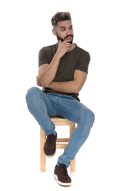 Pensive casual man looking away bothered with his hand on his chin while sitting on a chair on white studio background - Zdjęcie, obraz
