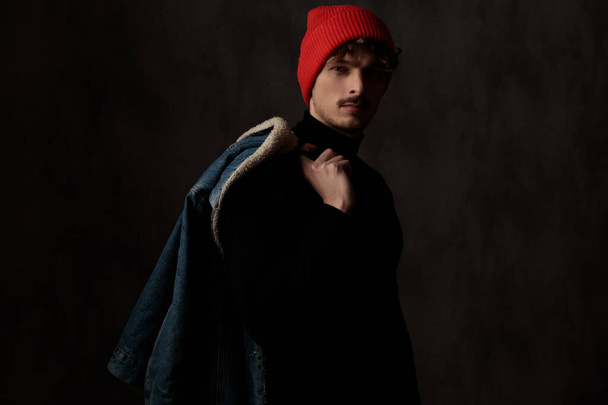 dramatic cool guy holding denim jacket over shoulders and posing on black background - Fotografie, Obrázek