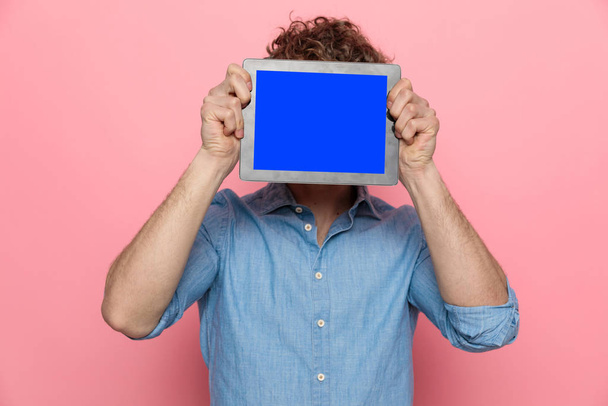 casual denim guy covering face with tablet and standing on pink background - Φωτογραφία, εικόνα