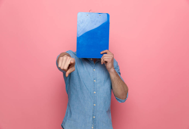 casual denim guy covering face with clipboard and pointing finger, standing on pink background - Fotoğraf, Görsel