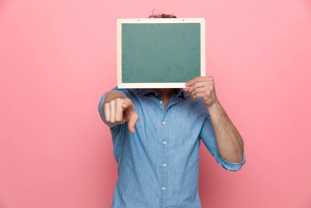casual guy hiding behind blackboard and pointing finger, standing on pink background - Foto, immagini