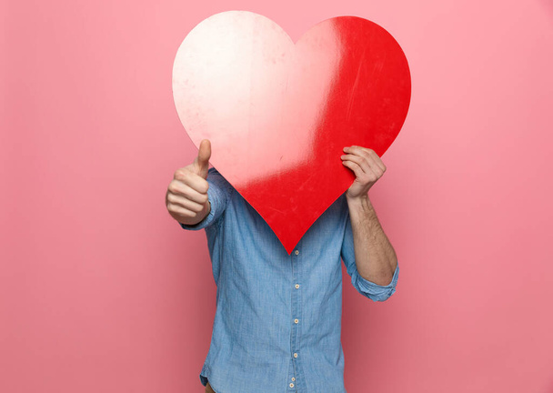 happy casual guy covering face with big red heart and making thumbs up sign, standing on pink background - Photo, image
