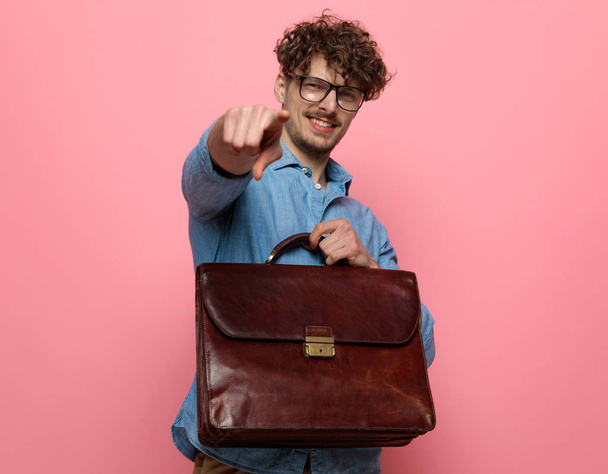 young casual man in denim shirt pointing finger and holding suitcase, standing on pink background - Φωτογραφία, εικόνα