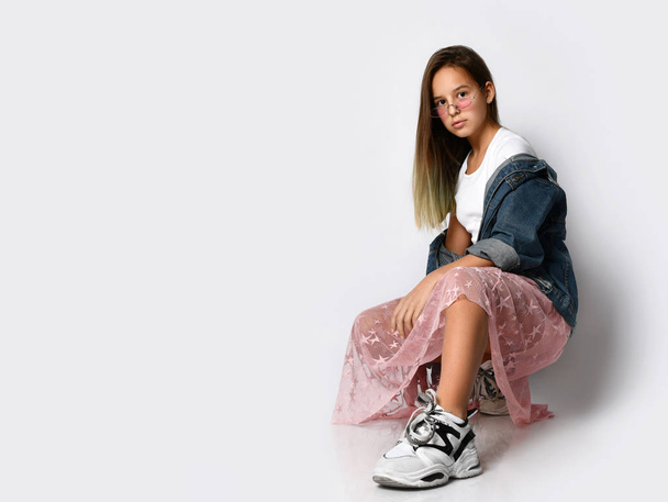 Young teen girl model in stylish casual clothes and sneakers sitting and looking at camera over white background - Fotó, kép