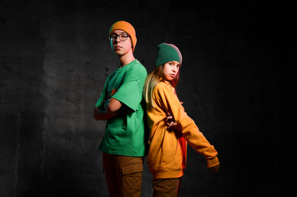 Young teens boy and girl in green and yellow stylish casual clothing and hats standing back to back over dark background - Fotó, kép